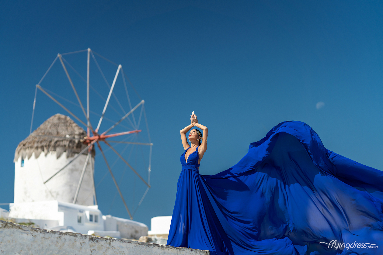 Woman in a royal blue flying dress with arms raised gracefully in front of the iconic Mykonos windmills, with the dress flowing beautifully against a vivid blue sky.
